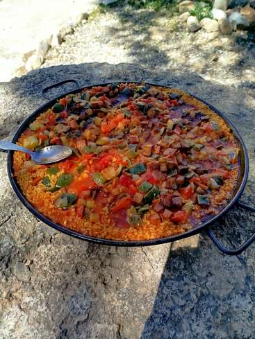 A large pan of colorful vegetable paella sits on a rustic stone table outdoors, filled with rice, zucchini, tomatoes, and peppers, with a serving spoon.