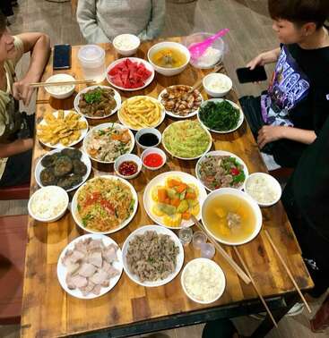 A group of three people sits around a wooden table filled with various Vietnamese dishes, rice bowls, soup, vegetables, fruit, and sauces, preparing to eat.