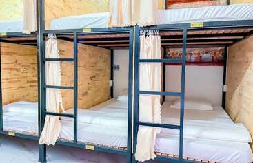 This image shows a set of modern bunk beds in a hostel dormitory. Each bed has white bedding, privacy curtains, wooden walls, and individual reading lights.