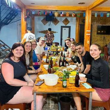 A group of friends enjoys a meal together at a long wooden table indoors. There are drinks, food, and international flags hanging above them. Smiling faces.