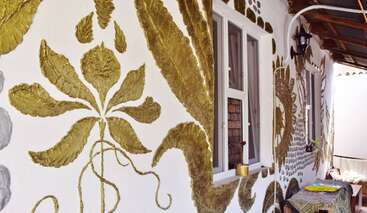Das Bild zeigt eine weiße Wand, die mit großen, reliefartigen goldenen Blumenmustern verziert ist. Zu sehen sind zwei Fenster, ein kleiner Tisch mit Gegenständen und eine Laterne.