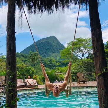 Um homem balança sobre uma piscina, cercado por uma vegetação exuberante e uma montanha à distância, desfrutando de um ambiente tropical sereno em um dia ensolarado.