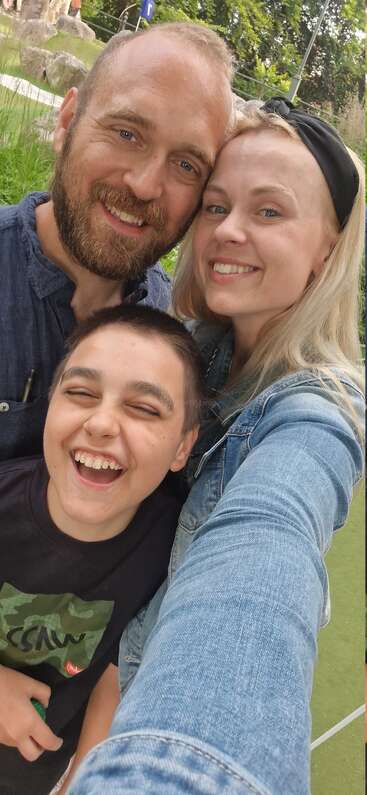 A joyful family selfie outdoors, featuring a smiling man, woman, and laughing boy. Everyone radiates happiness, enjoying a fun day together in a green park.