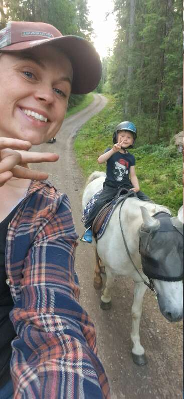 A smiling woman takes a selfie on a wooded trail. A child rides a white pony, both showing peace signs, surrounded by lush green forest.