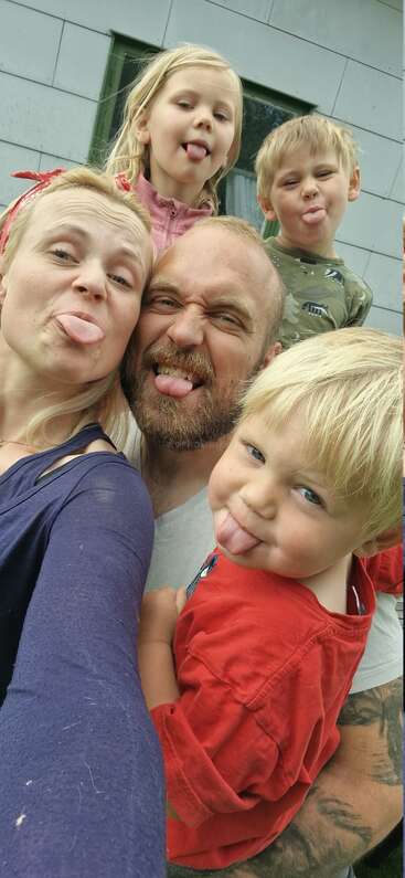 A joyful family group selfie outdoors, featuring two adults and three kids, all playfully sticking out their tongues and smiling at the camera. Happy, silly moment!