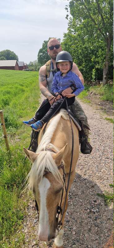 A man and smiling child ride a tan horse on a rural path, surrounded by green fields and trees, enjoying a sunny, peaceful countryside day together.