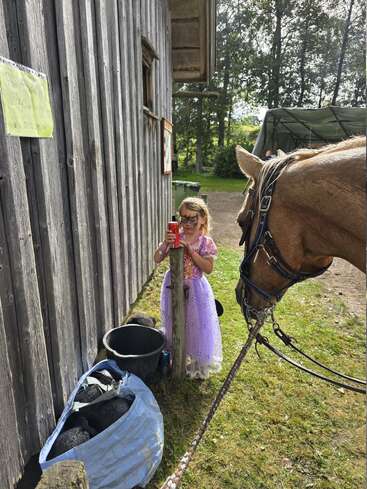 Uma jovem com um vestido roxo e pintura facial está ao lado de um cavalo do lado de fora de um celeiro de madeira, segurando uma xícara vermelha, em um dia ensolarado.