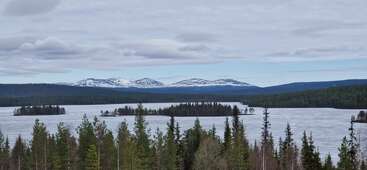 A serene, partially frozen lake surrounded by evergreen forests. Snow-capped mountains rise in the background under a cloudy sky. Small islands dot the tranquil water.
