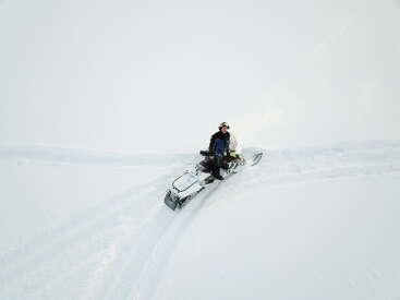A person rides a snowmobile across a vast, snowy landscape. The scene is minimalistic, with fresh tracks in the pristine white snow, evoking adventure.