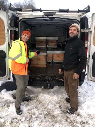 Deux personnes souriantes se tiennent près d'une camionnette enneigée dont les portes sont ouvertes, laissant apparaître plusieurs ruches en bois à l'intérieur. Elles semblent heureuses et sont habillées chaudement pour l'hiver.