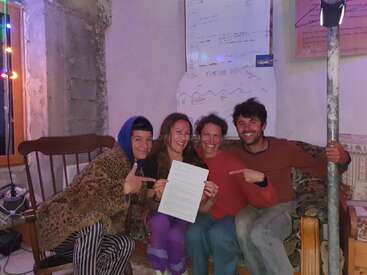 Quatre personnes souriantes sont assises sur un canapé à l'intérieur, tenant fièrement un document et le montrant du doigt. En arrière-plan, on aperçoit un tableau blanc avec des notes et des guirlandes lumineuses colorées.