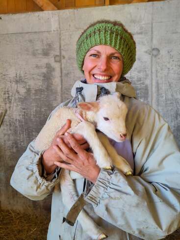 Une personne souriante, coiffée d'un bonnet vert en tricot et vêtue d'une veste grise, tient un petit agneau blanc dans une grange rustique aux murs en béton.