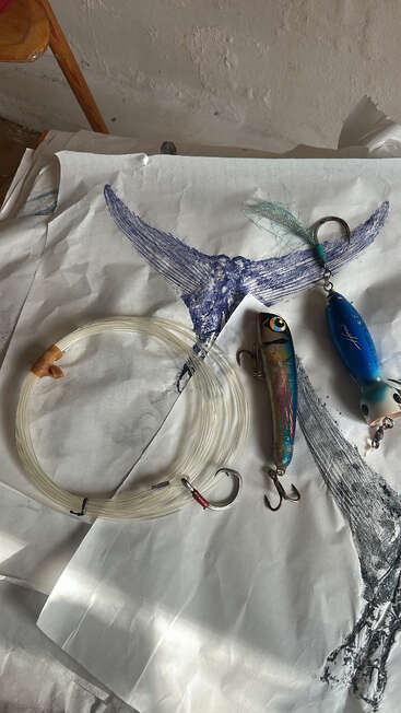 En esta imagen se ven señuelos de pesca, un sedal enrollado con anzuelos grandes y un papel con la huella de una cola de pez azul, dispuestos sobre una mesa.