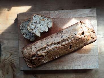 The image depicts a rustic wooden cutting board with a loaf of bread and two slices, set against a light-colored wood background with a subtle shadow cast by sunlight.