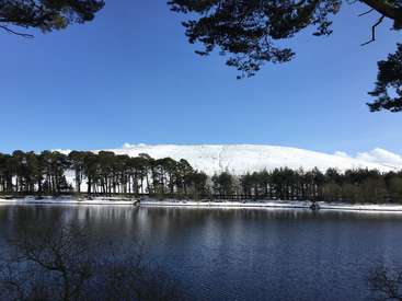 The image depicts a serene landscape featuring a snow-covered hill, a tranquil lake, and a forest of trees, set against a clear blue sky.