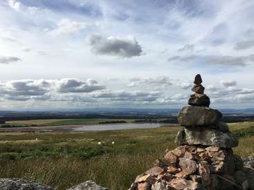The image depicts a serene landscape featuring a stack of rocks in the foreground, set against a backdrop of a vast field and a distant body of water under a cloudy sky.