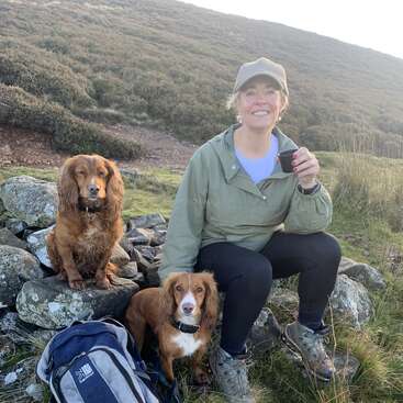 Uma mulher com roupas para atividades ao ar livre está sentada sobre pedras, sorrindo e segurando uma xícara. Dois cães marrons e uma mochila estão ao seu lado. Paisagem montanhosa e gramada ao fundo.