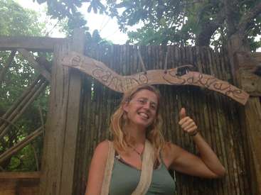 The image depicts a woman standing in front of a wooden fence with a sign that reads \"el camino salvaje\" in a natural setting, giving a thumbs up.