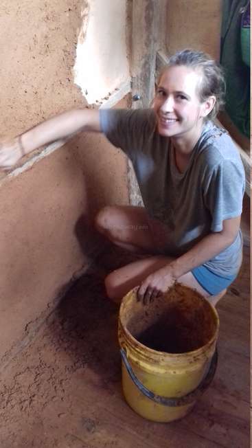 A woman kneels on the floor, smiling at the camera, with a bucket of mud beside her, applying mud to a wall in a rustic room.
