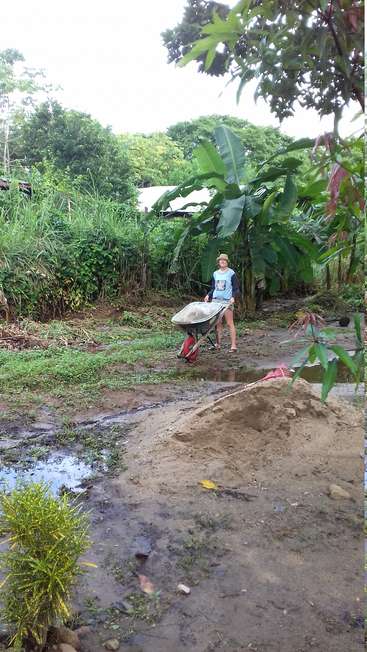 The image depicts a young girl standing in a muddy yard, pushing a wheelbarrow, surrounded by lush greenery and a small puddle of water.