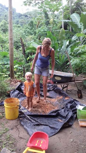A woman and a child stand in a pile of dirt on a tarp, surrounded by gardening tools and buckets in a lush, tropical environment.