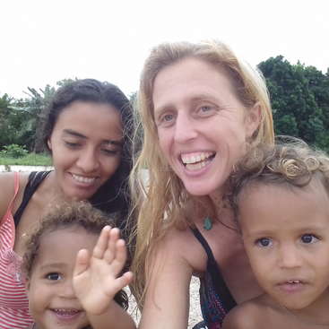 The image shows a woman with two children on a beach, with trees in the background and a cloudy sky. The woman has long blonde hair.