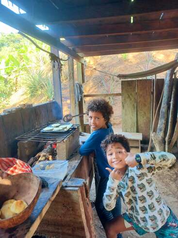 Two children smile and cook over a rustic outdoor wood stove. Surrounded by nature, they prepare food, enjoying the cozy, natural kitchen environment and each other's company.