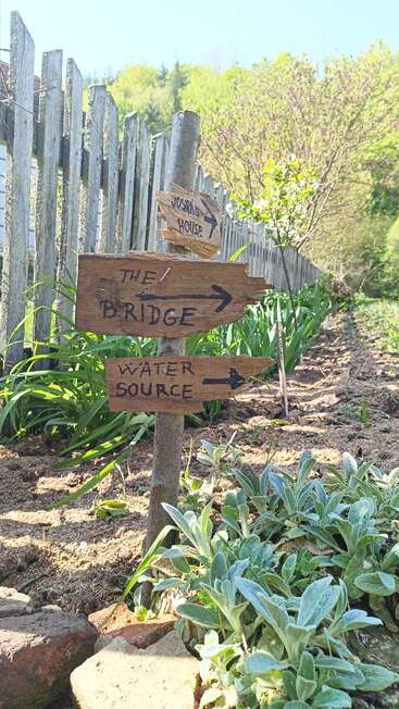 A rustic wooden signpost stands beside a weathered fence. Signs point towards the Josiah House, the bridge, and a water source amidst a lush garden.