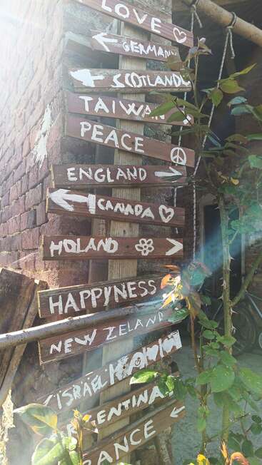Wooden signposts painted with country names (Germany, Scotland, Taiwan, etc.) and words like "LOVE," "PEACE," and "HAPPINESS" beside a rustic brick wall.