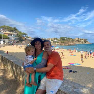La imagen muestra a una familia de tres miembros posando para una foto en un muro de piedra con vistas a una playa de arena con un océano de un azul vibrante y una pintoresca ciudad al fondo.