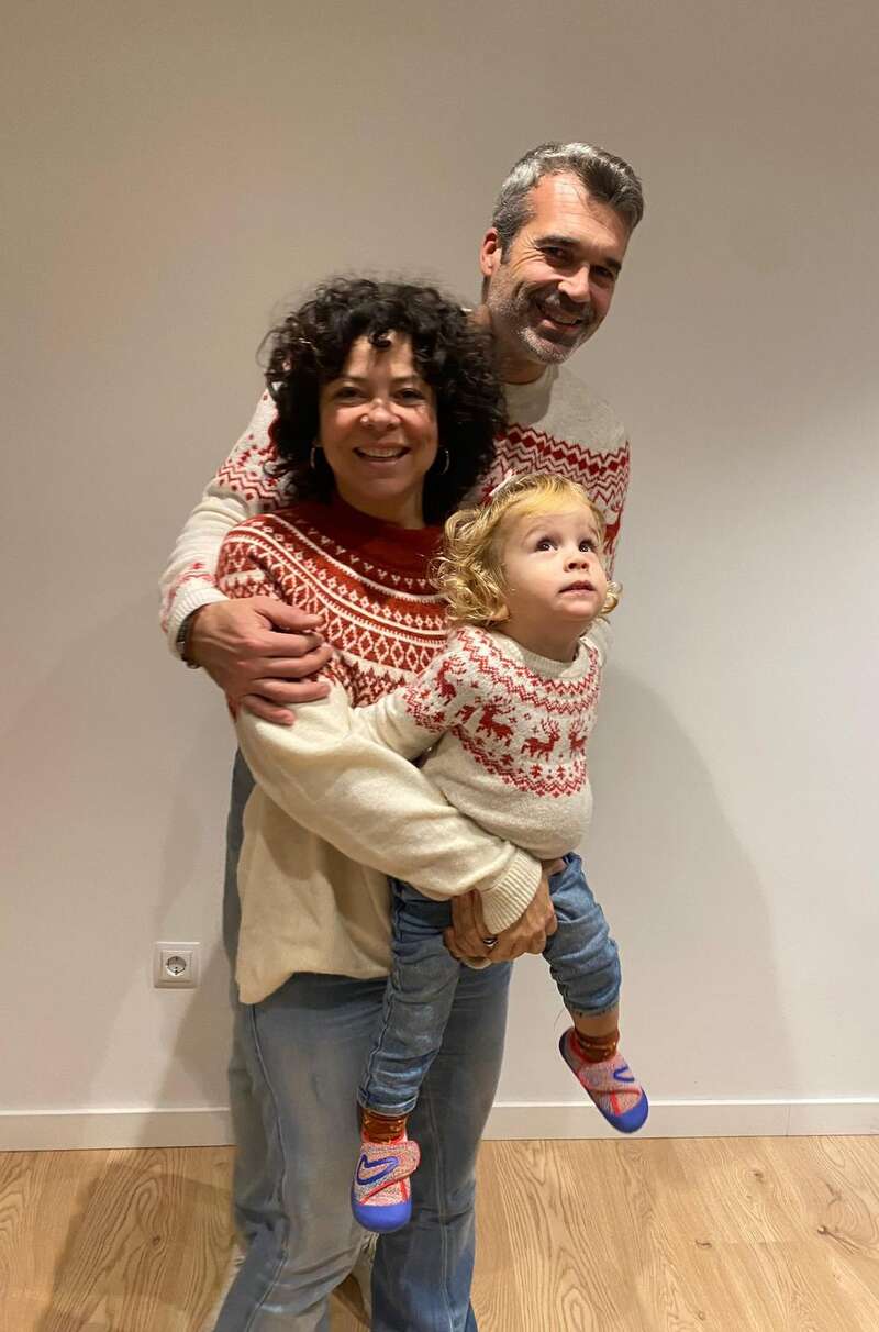 A imagem mostra uma família de três pessoas posando para uma foto, com os pais abraçando o filho pequeno, todos vestindo suéteres vermelhos e brancos combinando.