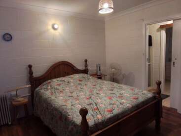 Une chambre confortable comprend un lit en bois avec une couette à motifs floraux, des tables de chevet, une lampe, un ventilateur, des murs en briques blanches, une horloge et une salle de bains attenante.