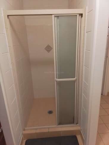 Esta imagen muestra una pequeña cabina de ducha alicatada con una puerta corredera de cristal esmerilado, baldosas beige en el suelo y un único azulejo decorativo en la pared trasera.