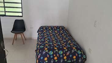 Un dormitorio sencillo y despejado, con paredes blancas, suelo de baldosas y una cama individual cubierta con una manta colorida con motivos espaciales. Junto a la ventana hay una silla negra.