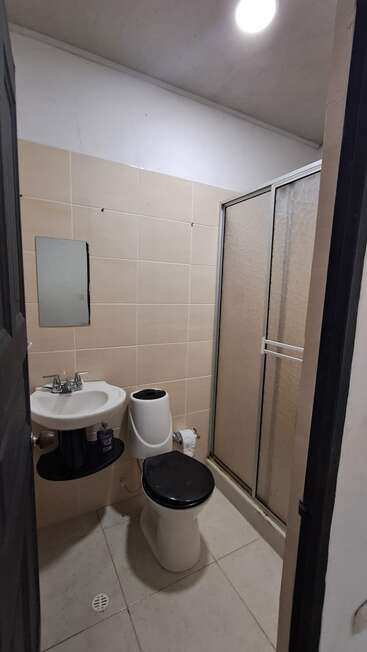 Este pequeño cuarto de baño cuenta con paredes alicatadas en tonos beige, un lavabo blanco con espejo, un inodoro con asiento negro y una mampara de ducha corredera de cristal.