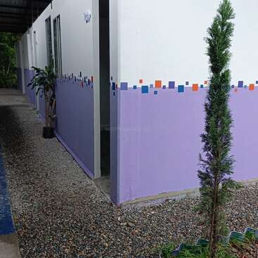 Una esquina de un edificio con paredes de color morado claro, decoradas con pequeños cuadrados de colores. En el exterior, junto al edificio, hay macetas con plantas y un camino de grava.
