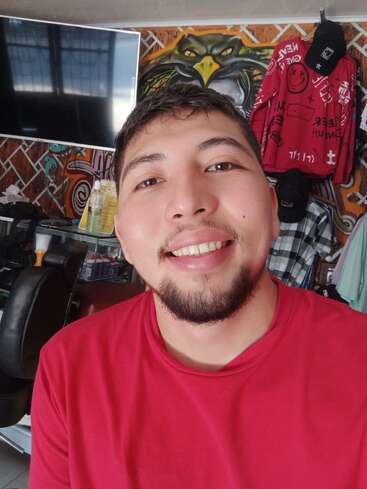 Un joven con barba sonríe para hacerse un selfi en una habitación con grafitis de colores, un televisor, una silla de barbero, camisetas y un llamativo mural de un águila.
