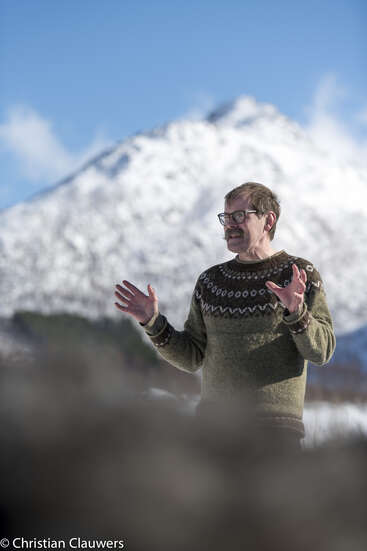 Ein Mann mit Brille und einem gemusterten grünen Pullover gestikuliert mit den Händen, während er draußen steht, im Hintergrund ein schneebedeckter Berg und ein blauer Himmel.
