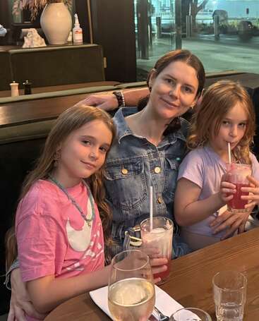 Une femme est assise à la terrasse d'un restaurant avec deux jeunes filles, toutes souriantes et dégustant ensemble des boissons roses. L'atmosphère est chaleureuse et détendue.