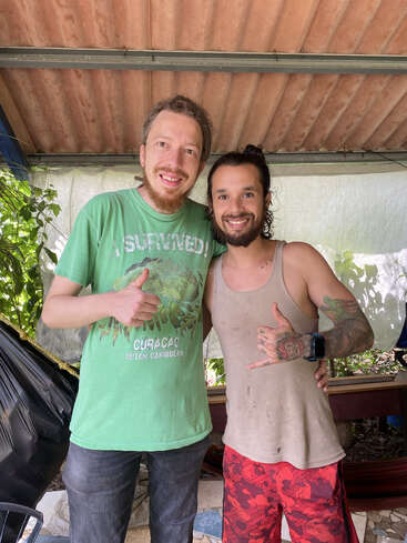 Dois homens sorridentes lado a lado, um de camisa verde e jeans, o outro de camiseta regata e bermuda vermelha, posando com gestos amigáveis.