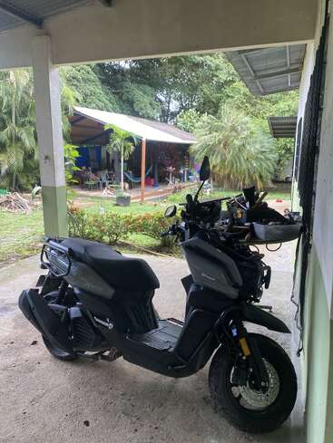Uma scooter preta está estacionada em uma área coberta ao lado de uma casa. Ao fundo, há um pátio com cadeiras, plantas e um ambiente verde exuberante.