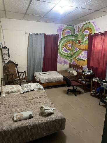 Um quarto aconchegante com duas camas, uma cadeira de balanço, uma escrivaninha, sapatos e prateleiras. Um grande mural de dragões coloridos decora a parede do canto, acrescentando personalidade.