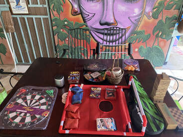 Una mesa está llena de juegos de mesa, cartas, un tablero de dardos, Jenga, pufs y material de ping pong, con un vibrante mural de una cara morada como fondo.