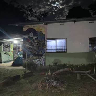 Uma cena noturna mostra um prédio com um mural colorido, barracas montadas sob uma varanda, troncos ao redor de uma fogueira e um céu nublado e iluminado pela lua acima.