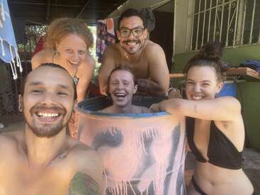 Cinco amigos sonríen y ríen juntos al aire libre. Una persona está sentada dentro de un barril pintado, mientras que los demás se reúnen a su alrededor, disfrutando de un momento divertido.