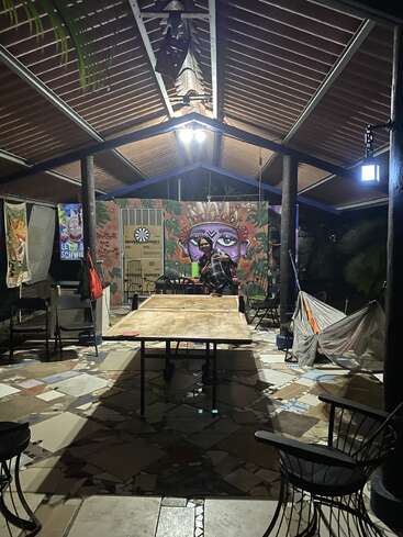 Un pabellón al aire libre por la noche con una mesa de ping pong, un mural colorido, un tablero de dardos, sillas, hamacas y una persona de pie cerca de la parte trasera, iluminada por luces cenitales.