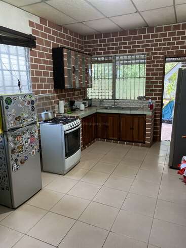 Cette cuisine est dotée de murs en briques, d'un sol carrelé, d'un réfrigérateur recouvert d'autocollants, d'une cuisinière, d'armoires en bois et de fenêtres avec des grilles métalliques, créant ainsi un espace confortable et chaleureux.