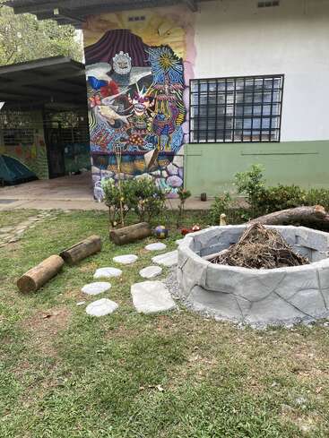 Une fresque colorée orne le mur d'un bâtiment derrière un jardin agrémenté d'un foyer circulaire en pierre, de sièges en rondins, de dalles et d'une pelouse verte. Cadre extérieur artistique.