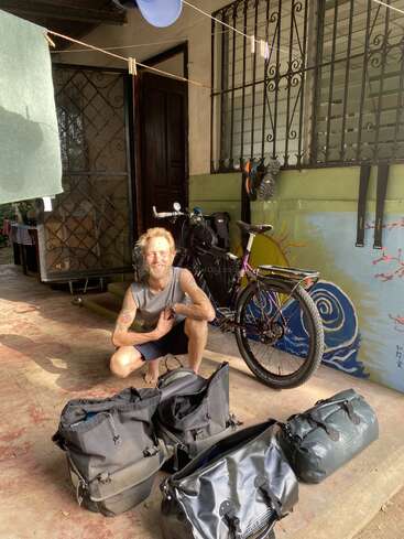Un homme au bras tatoué est agenouillé près d'un vélo chargé, entouré de grands sacs, sous un porche ombragé aux murs peints et aux barreaux de fer.