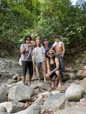 Cinq amis, pieds nus, se tiennent debout et accroupis sur de gros rochers dans une forêt tropicale luxuriante. Ils sourient à la caméra, profitant ensemble d'une aventure en plein air.